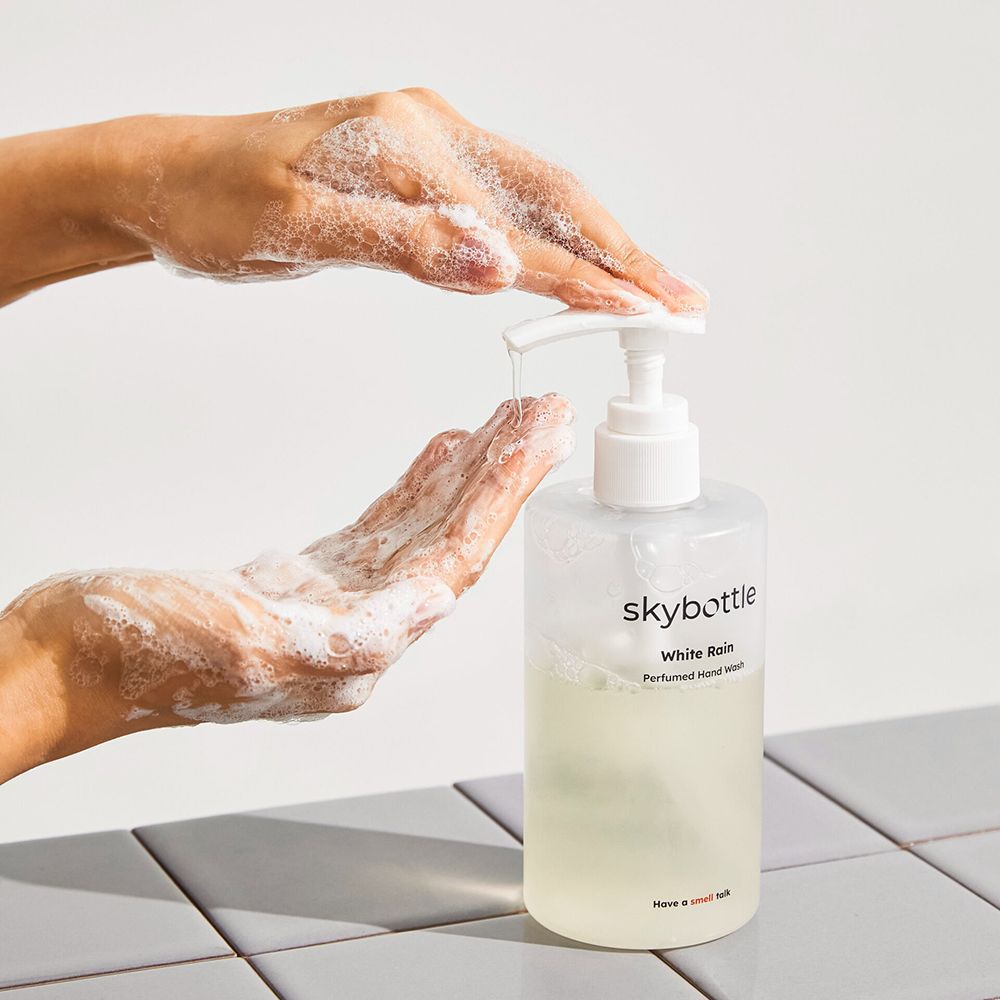 Hände, die Flüssigseife aus einer Flasche mit weißem Pumpverschluss entnehmen. Aufschrift: skybottle, White Rain, Perfumed Hand Wash.