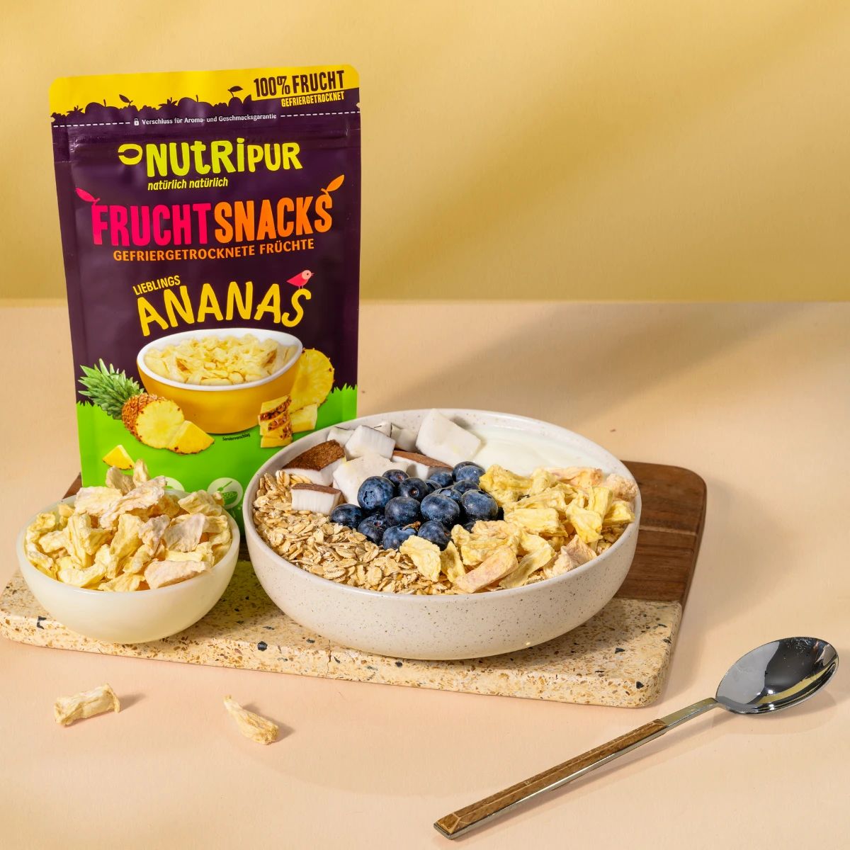 Beutel mit gefriergetrockneten Ananas-Bits. Daneben eine Schale mit Müsli, Blaubeeren und Kokosraspeln. Aufschrift: NutriPur, Fruchtsnacks.