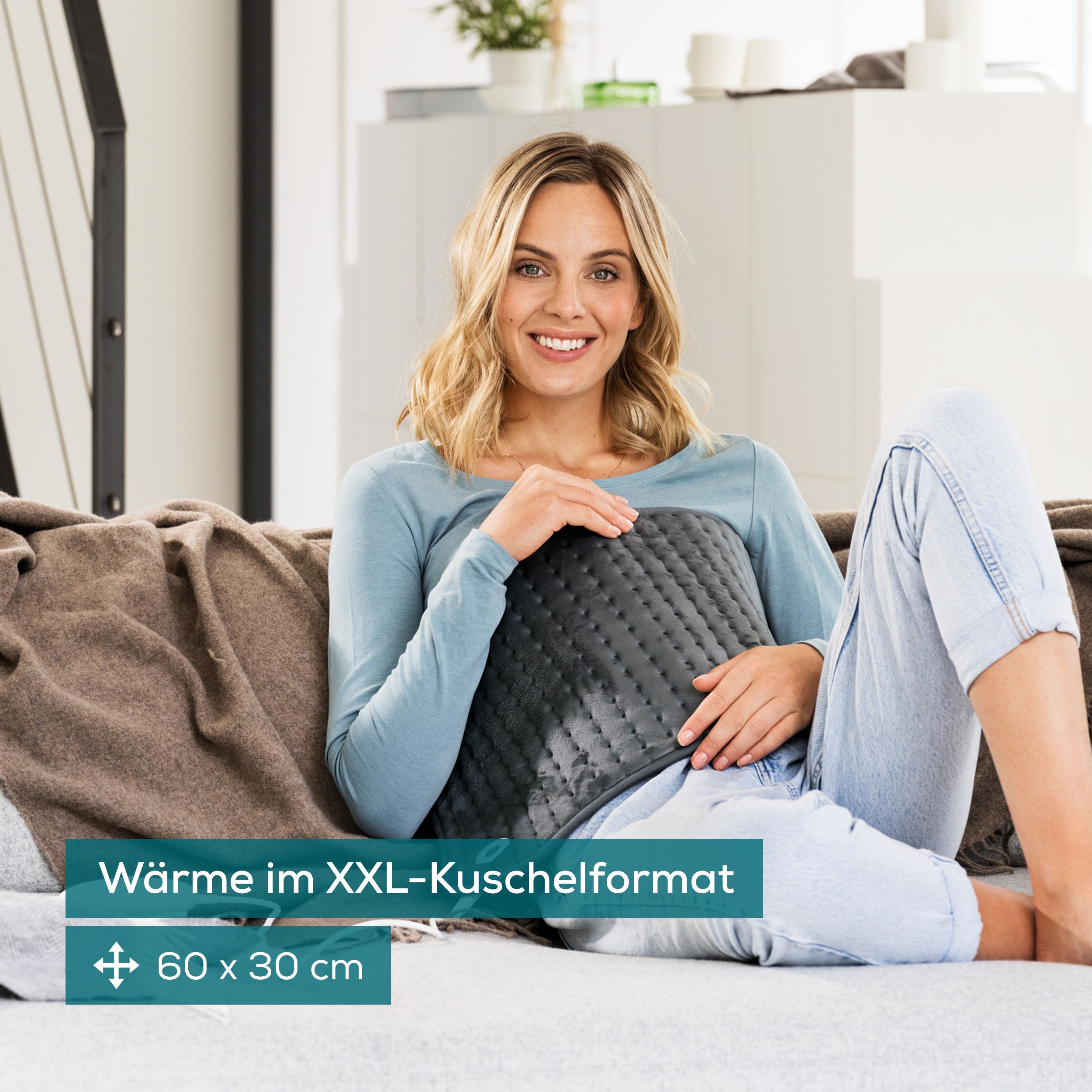 Frau sitzt auf Sofa, Heizkissen auf dem Schoß. Graues Heizkissen mit Steuerung. Text: Wärme im XXL-Kuschelformat. Maße: 60 x 30 cm.