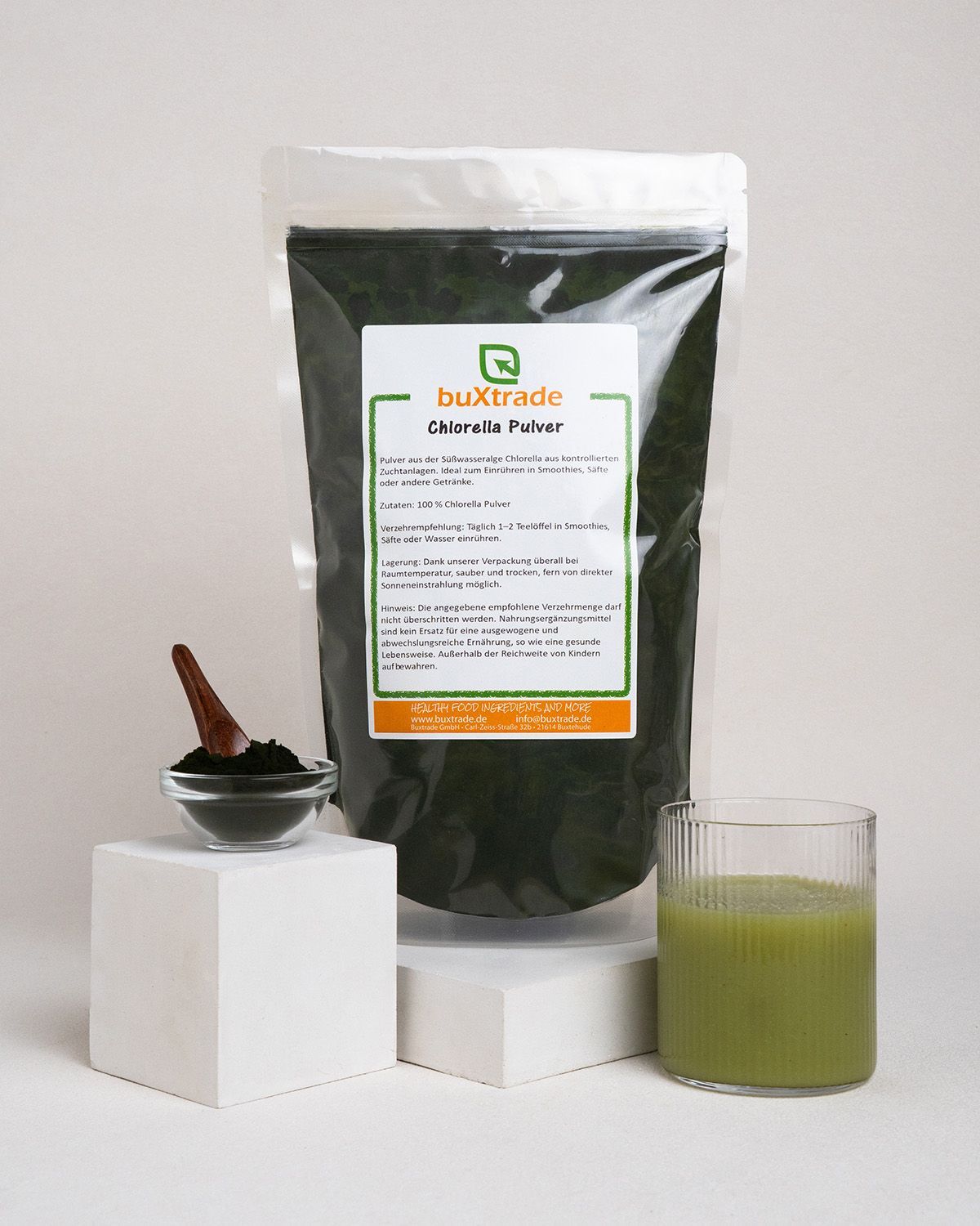 Glas mit grünem Getränk, Schale mit Pulver, Holzlöffel und Beutel mit buXtrade Chlorella Pulver. Auf Podest.
