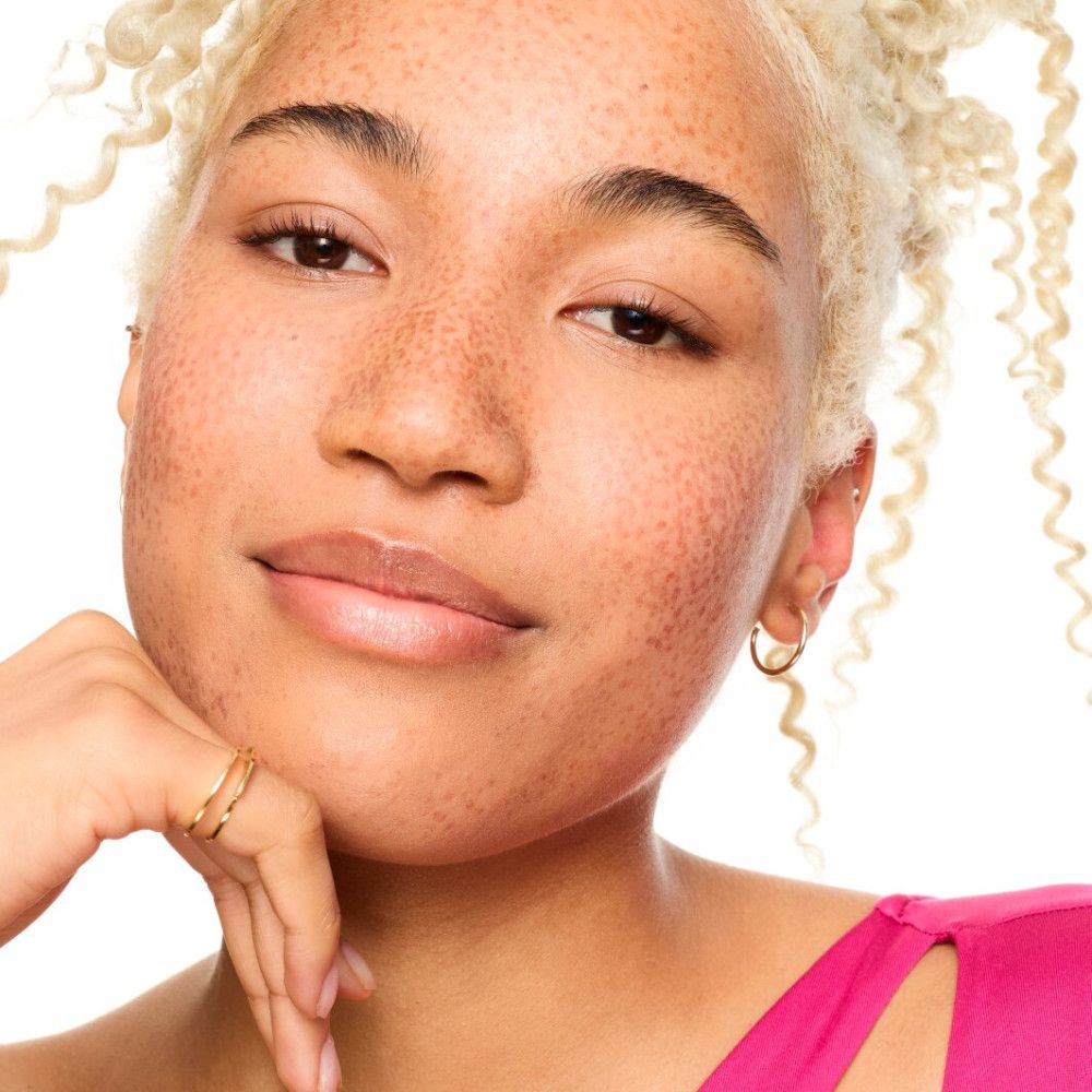 Nahaufnahme einer Frau mit Sommersprossen. Sie berührt ihr Kinn. Sie trägt Ohrringe und ein pinkes Oberteil.