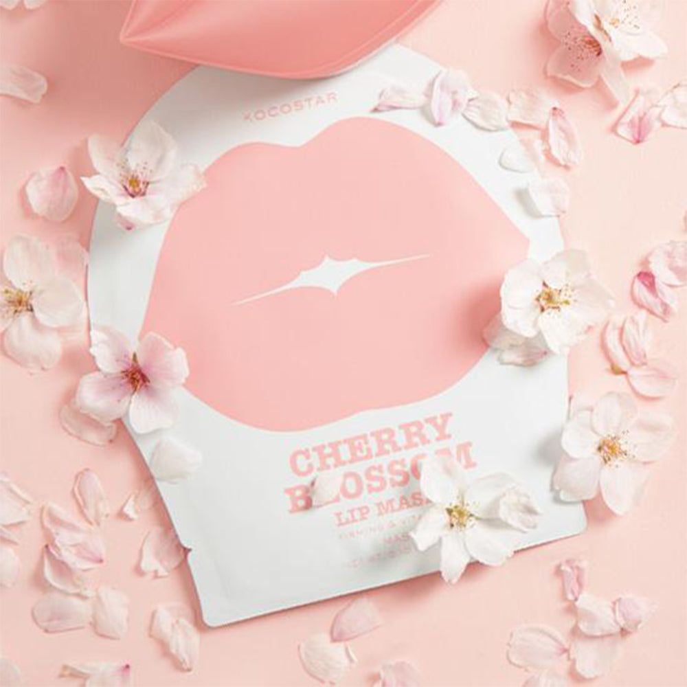 Verpackung mit Lippenmaske, umgeben von Blütenblättern und Blüten. Rosa Hintergrund. Text: Cherry Blossom Lip Mask. Marke: KOCOSTAR.