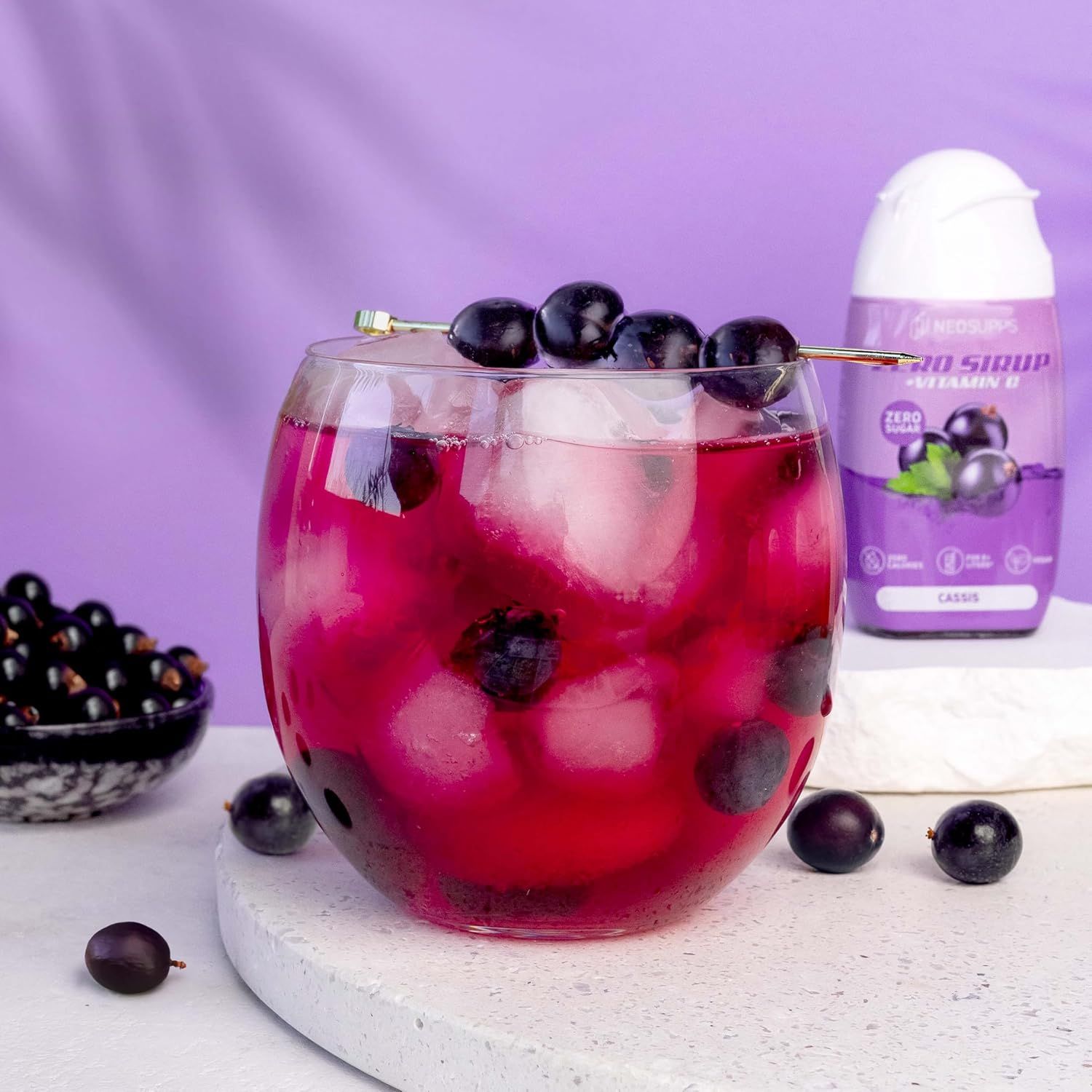Glas mit Getränk, Eiswürfeln und Beeren. Flasche mit Produkt daneben. Aufschrift: Zero Sirup, Cassis.