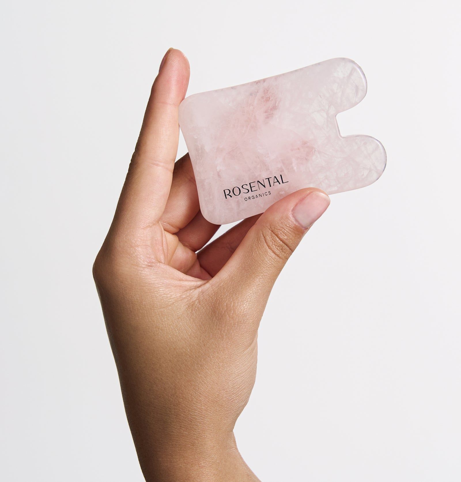 Gua Sha aus Rosenquarz, gehalten von einer Hand. Rosa Stein, abgerundete Form mit zwei Aussparungen. Text: Rosental Organics.