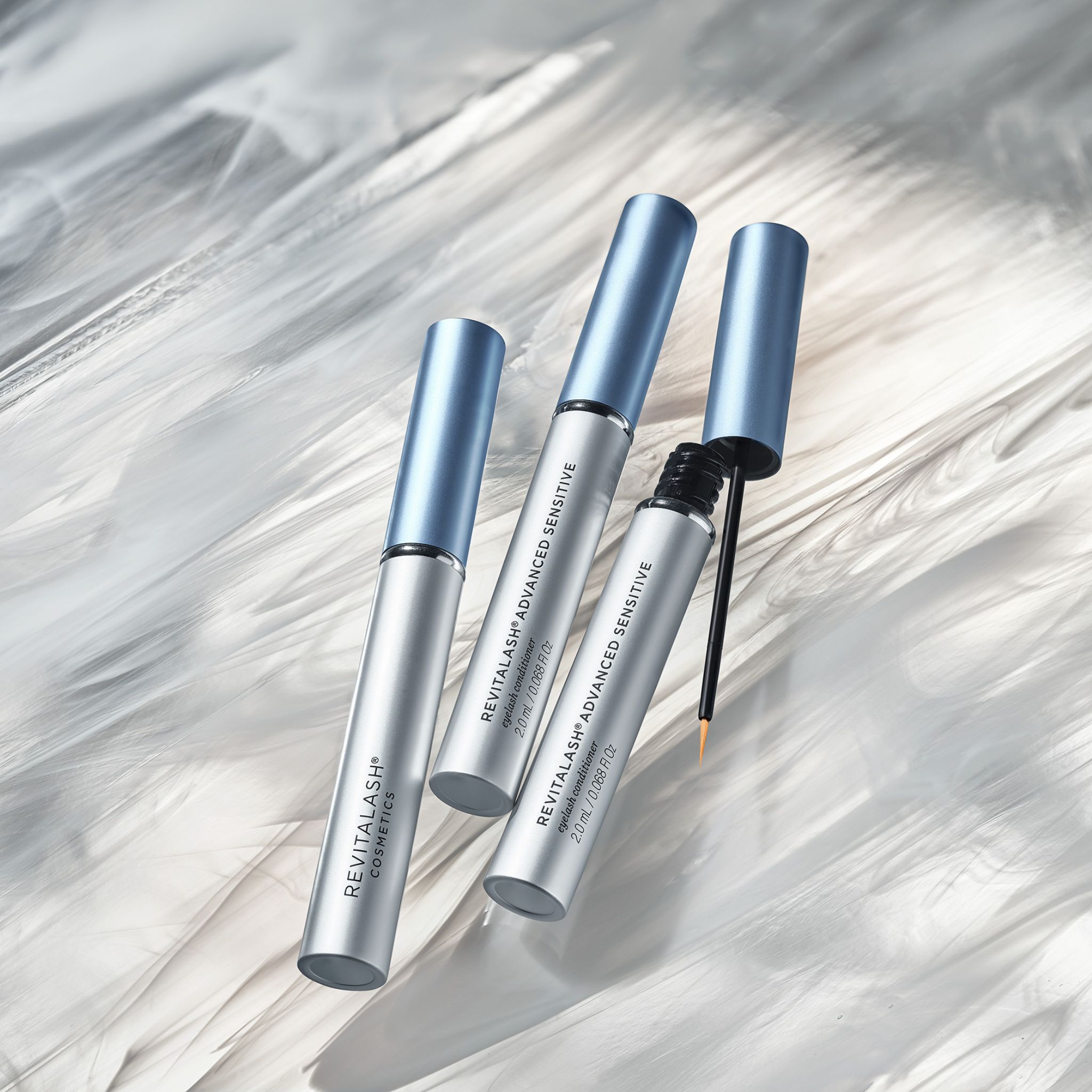 Drei Flakons RevitaLash® Advanced Sensitive, einer mit geöffnetem Applikator, auf weißem Hintergrund.