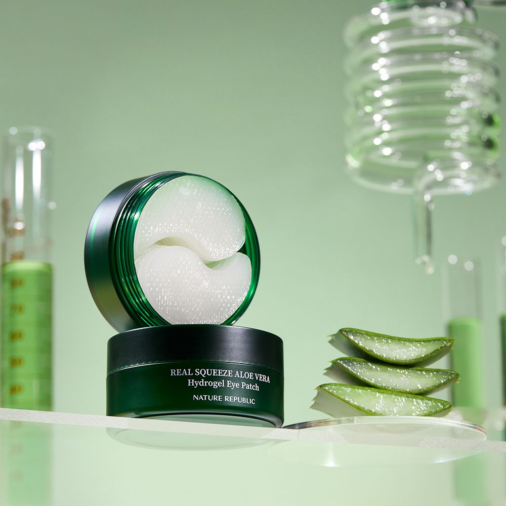Geöffnete grüne Dose mit Augen-Pads. Daneben Aloe-Vera-Blätter. Im Hintergrund Laborutensilien.
