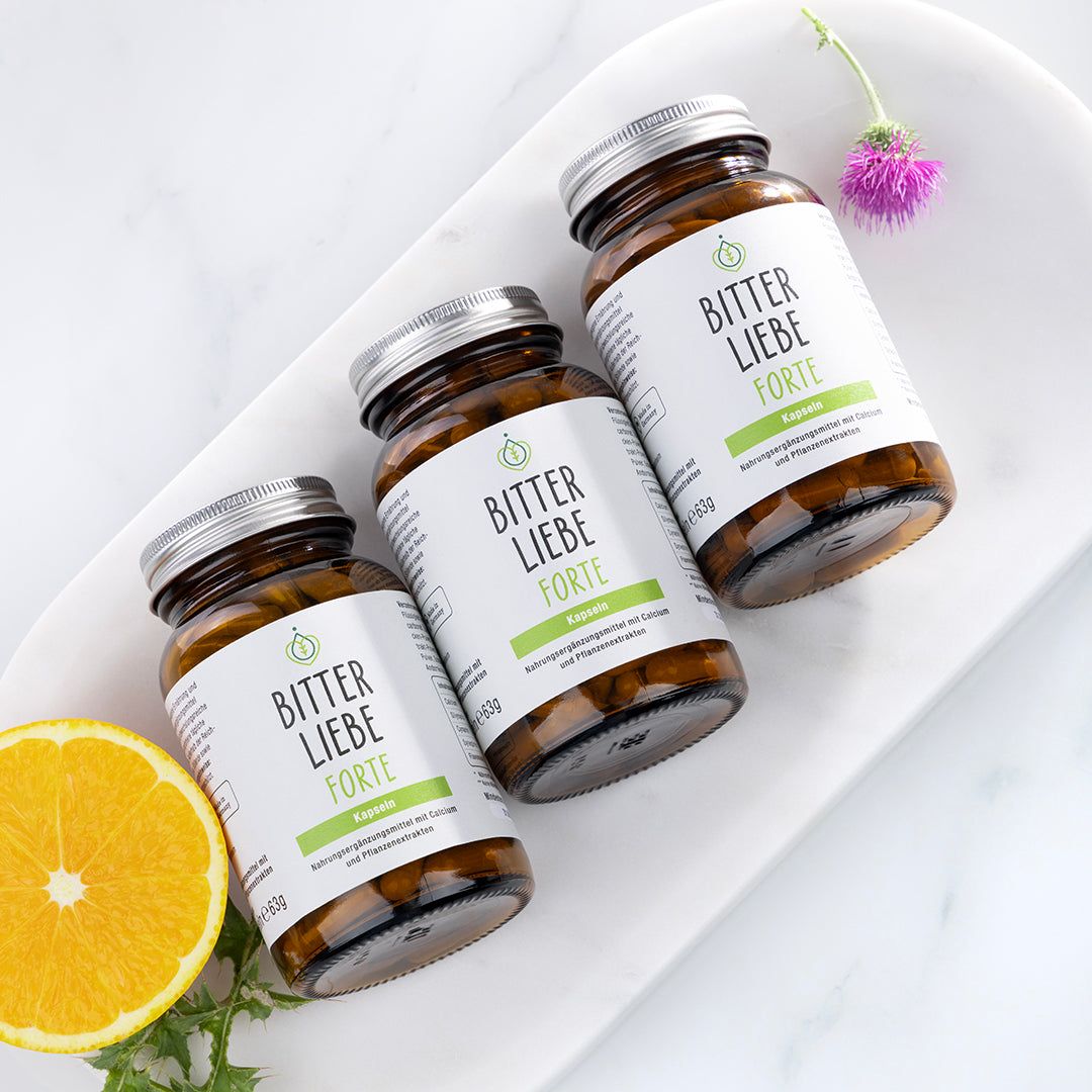 Drei Flaschen "BitterLiebe Forte" auf weißer Platte. Flaschen mit silbernen Deckeln und weißen Etiketten.