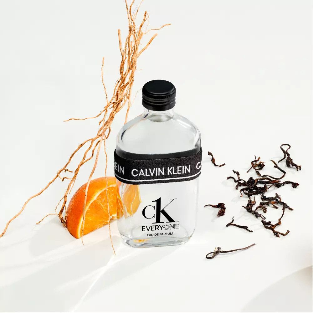 Klare Glasflasche mit schwarzem Deckel und Band. Aufschrift: Calvin Klein, ck EVERYONE. Mit Orange und Zweigen.