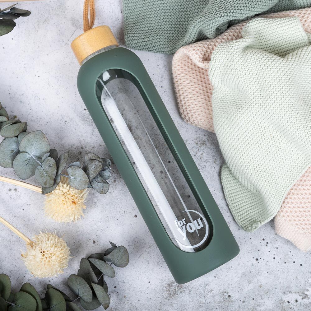 Grüne Glas-Trinkflasche mit Bambusdeckel, "for you" Logo, umgeben von Trockenblumen.