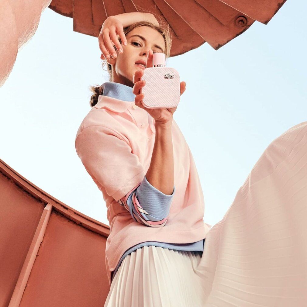 Frau hält rosa Lacoste L.12.12 Rose EdP Nat. Spray Flakon. Sie trägt rosa Oberteil und weißen Rock. Hintergrund: rotes Geländer und blauer Himmel.