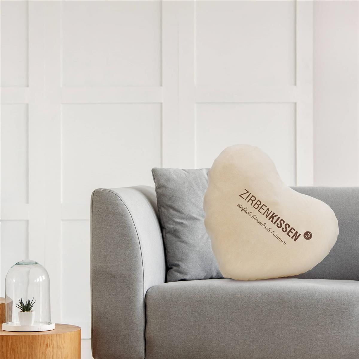 Beiges, herzförmiges Kissen auf einer Couch. Text: Zirbenkissen. Aufschrift: einfach himmlisch träumen. Kissen auf Couch.
