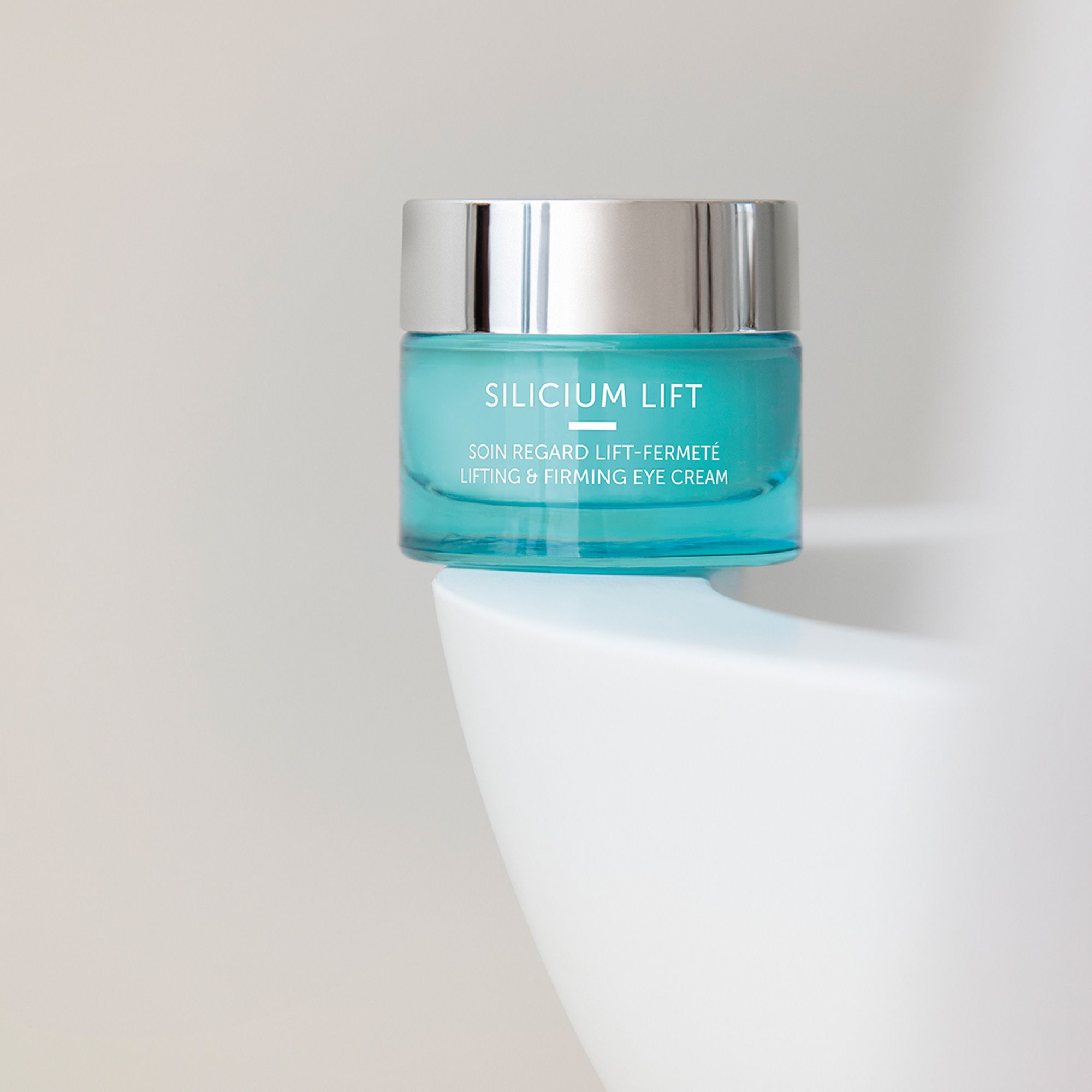 Türkisfarbener Tiegel mit silbernem Deckel. Aufschrift SILICIUM LIFT, darunter SOIN REGARD LIFT-FERMETÉ und LIFTING & FIRMING EYE CREAM. Auf weißer Oberfläche.