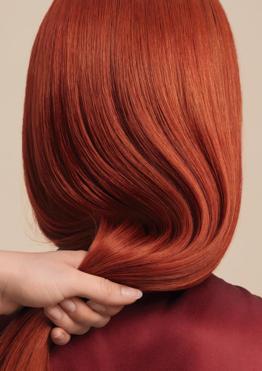 Rotes Haar, Hand hält Haarsträhne. Hintergrund beige. Person trägt rotes Oberteil.