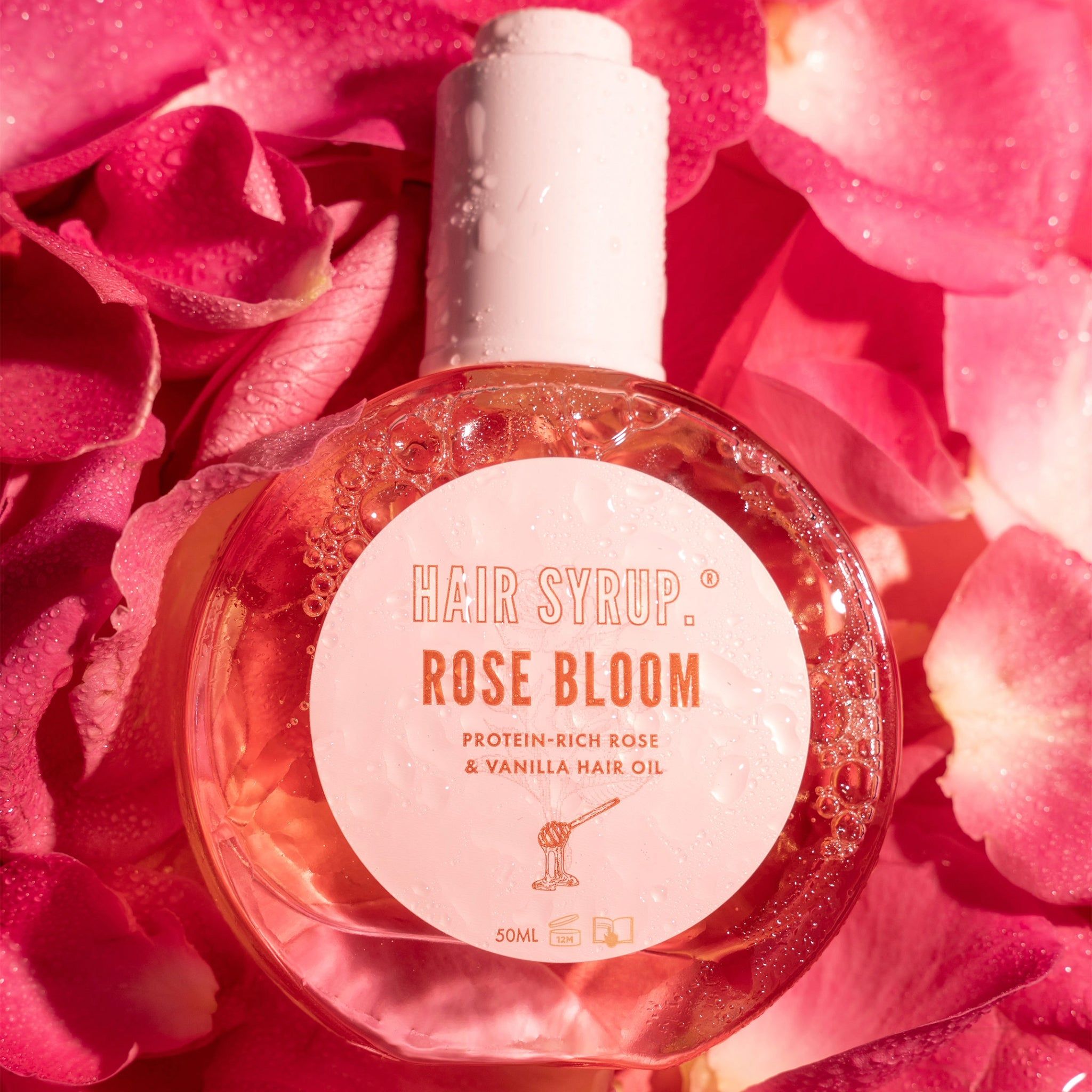 Runde Flasche mit weißem Verschluss, umgeben von Rosenblättern. Aufschrift: Hair Syrup Rose Bloom. Proteinreiches Rosen- und Vanille-Haaröl.