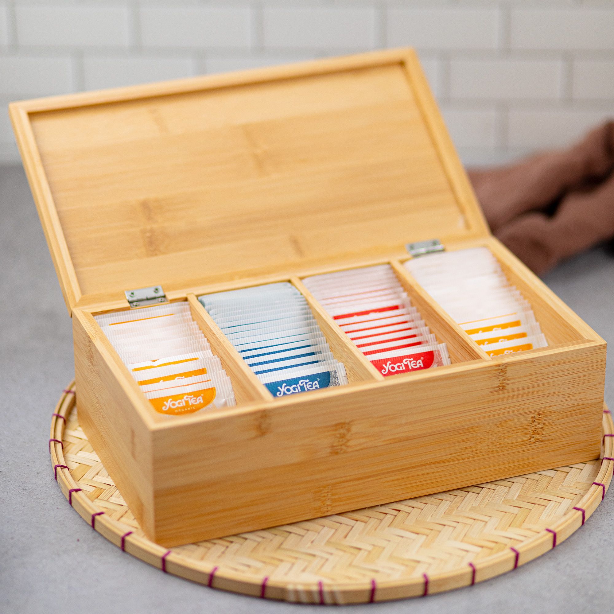 Geöffnete Holzbox mit vier Fächern, gefüllt mit Teebeuteln. Die Box steht auf einem geflochtenen Untersetzer.