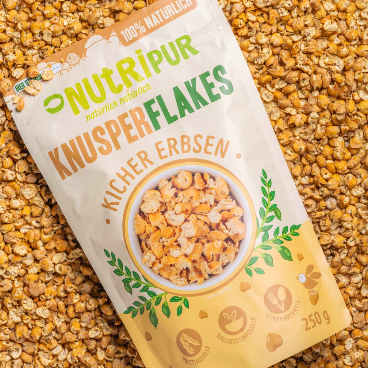 Nahaufnahme von Knusperflakes Kichererbsen. Die Flakes sind hellbraun und liegen dicht an dicht. Die Packung Knusperflakes Kichererbsen ist im Hintergrund zu sehen.