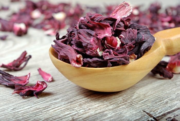 Holzlöffel mit getrockneten Hibiskusblüten. Rote Blütenblätter. Auf hellem Holzuntergrund.