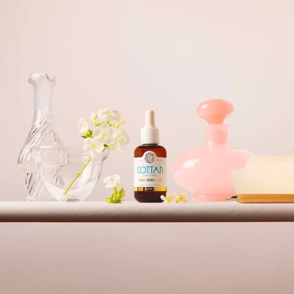 Serumflasche mit Tropfer, daneben Glasvasen und eine rosa Flasche. Aufschrift: COTTAN, Serum Bonne Mine.
