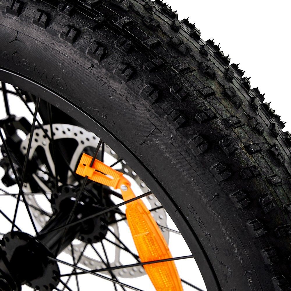 Nahaufnahme eines schwarzen Fahrradreifens mit grobem Profil. Scheibenbremse und Speichenrad sichtbar. Orangefarbener Reflektor.