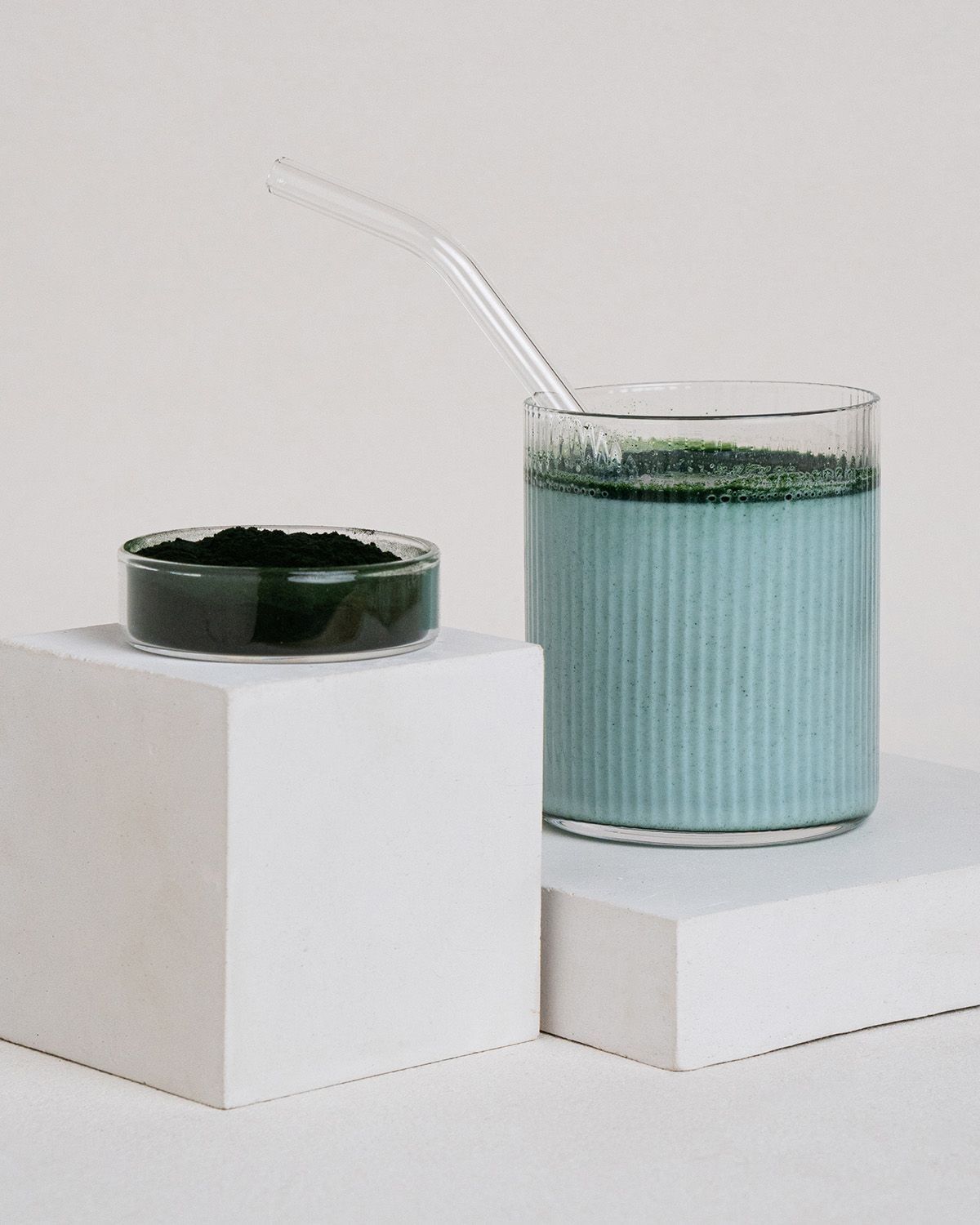 Glas mit blauem Getränk und Spirulina-Pulver. Ein Glasstrohhalm steckt im Getränk. Kleines Schälchen mit Pulver auf weißem Podest.