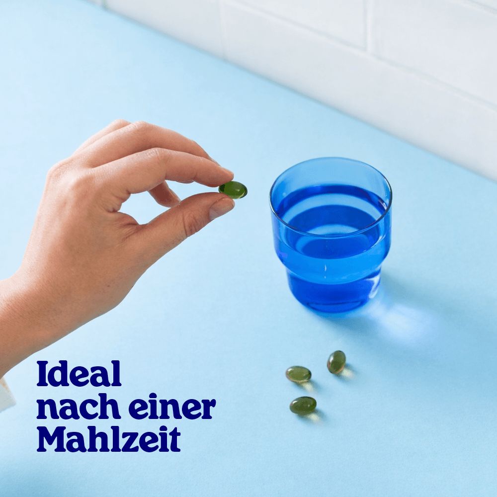 Grüne Kapsel in Fingerhaltung, neben einem blauen Glas und drei weiteren Kapseln.