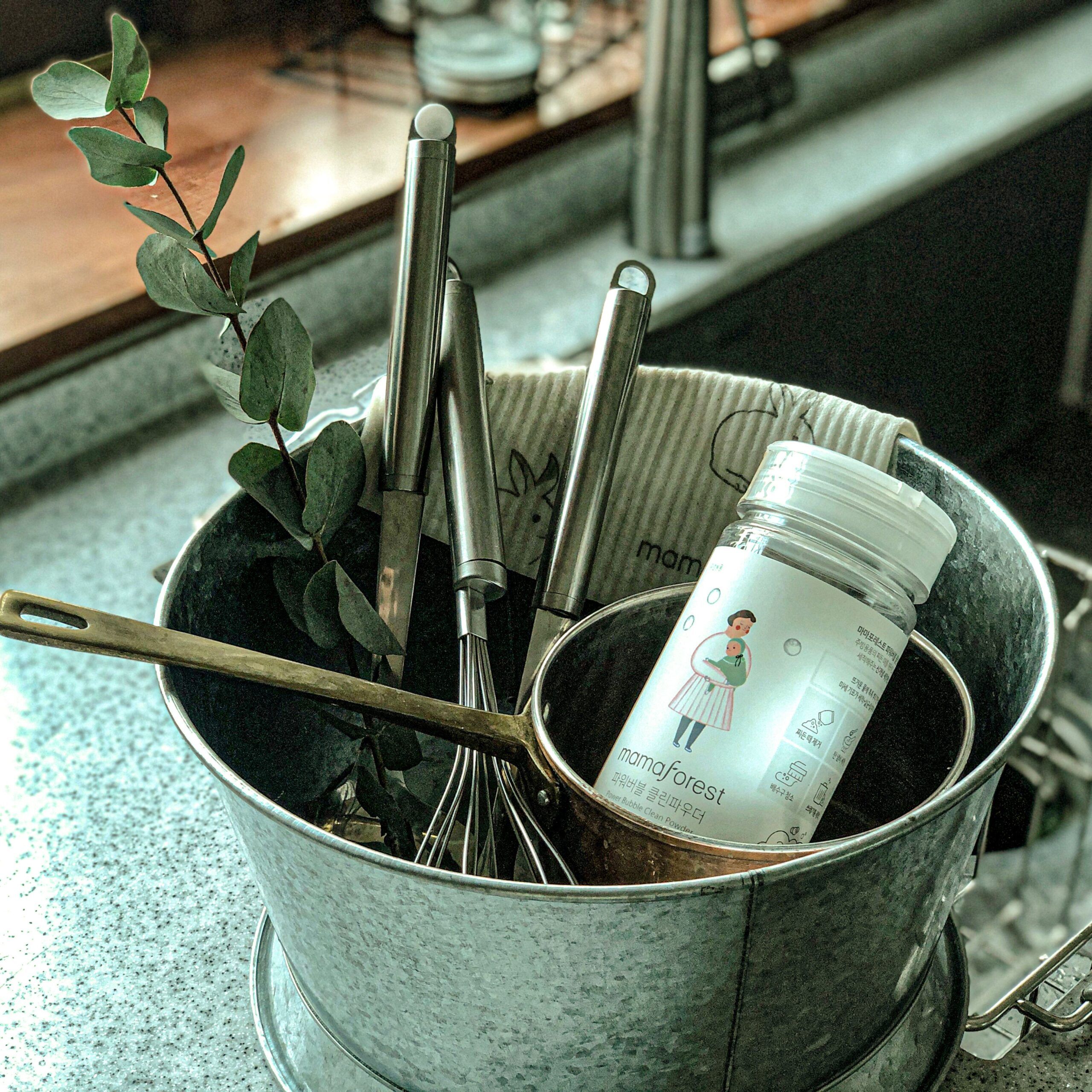 Metallbehälter mit Küchenutensilien und Flasche "Mighty Bubble Powder". Flasche mit Etikett. Hintergrund: Spüle und Fenster.