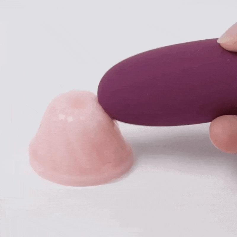 Lila Wellness-Produkt berührt eine rosa Substanz. Finger halten das Produkt. Helle Oberfläche.