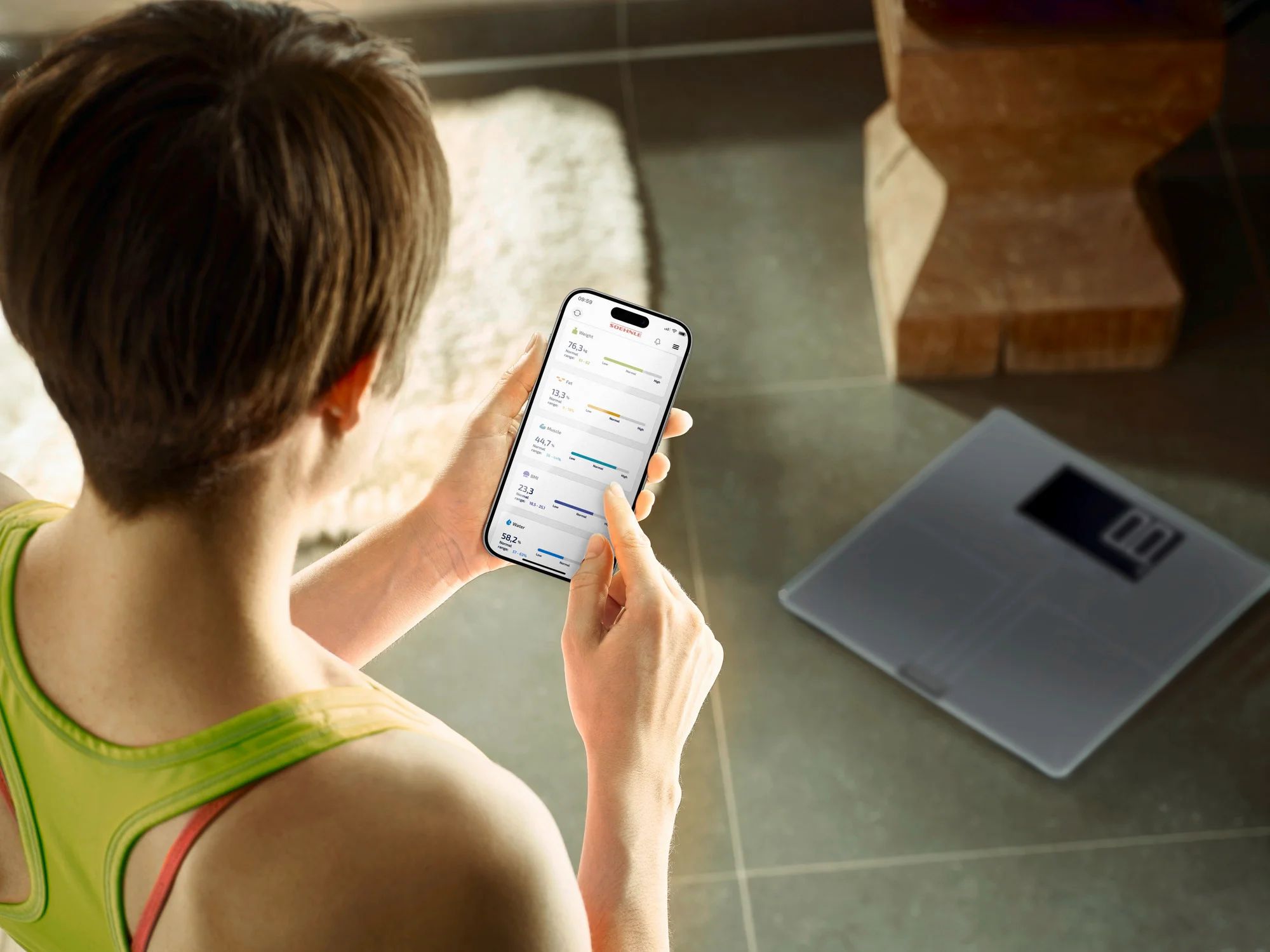 Frau betrachtet Smartphone mit Gesundheitsdaten. Daneben eine digitale Waage. Badezimmerumgebung.