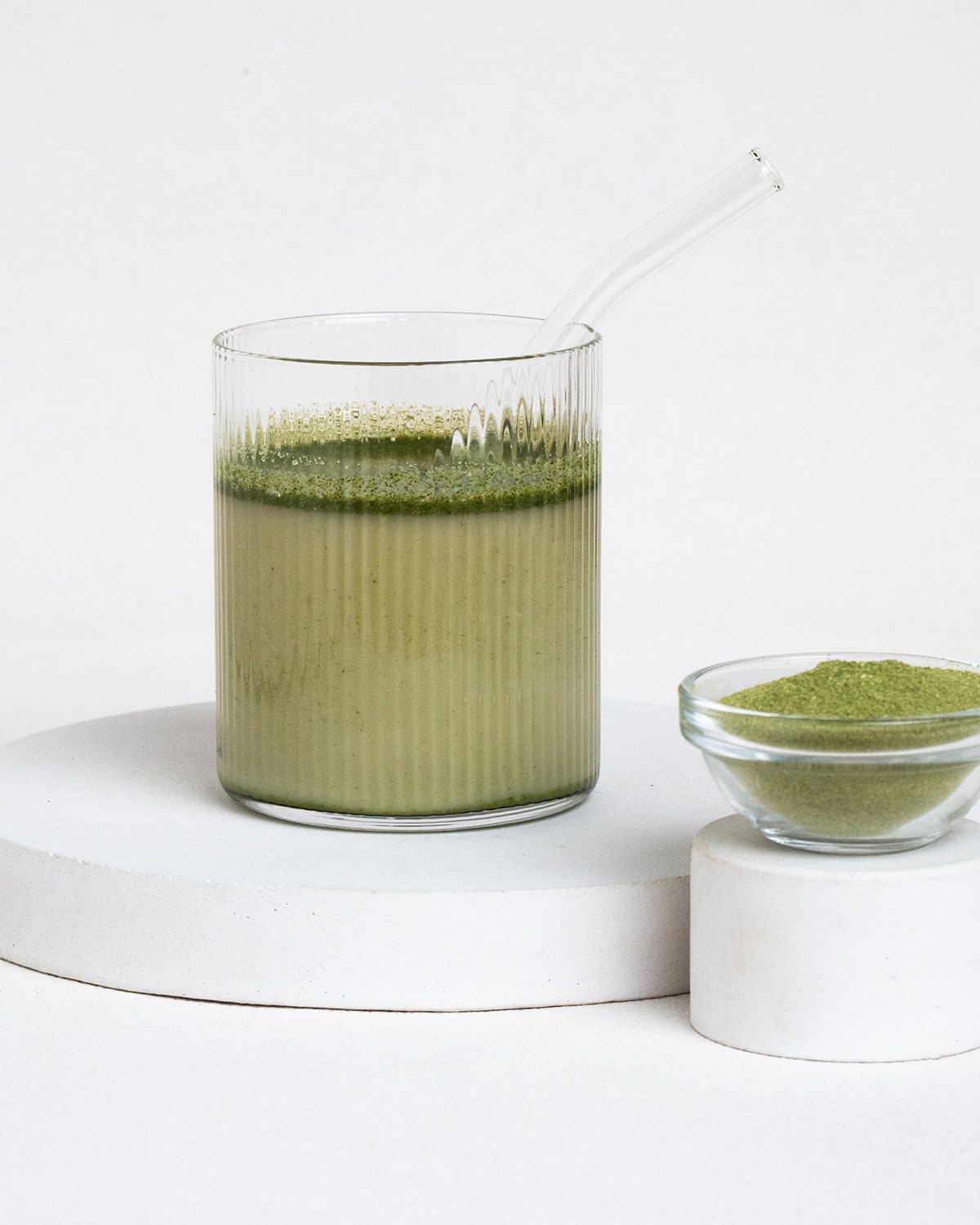 Glas mit grünem Getränk und Schale mit Moringa-Pulver. Mit Trinkhalm. Auf weißem Podest.