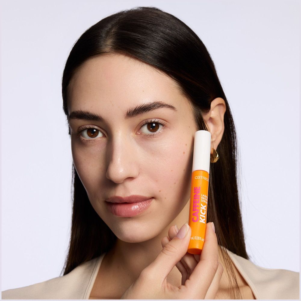 Frau hält das orangefarbene Serum. Aufschrift: Caffeine Kick, Lash & Brow Serum. Auf dem Deckel: Proven Fuller Lashes & Brows in 4 Weeks. Helle Haut.