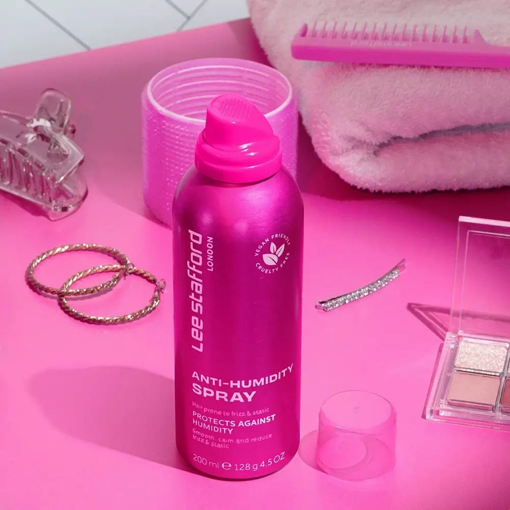Rosa Sprühflasche Lee Stafford Anti-Humidity Spray. Vegan, tierversuchsfrei. Auf rosa Untergrund mit Accessoires.