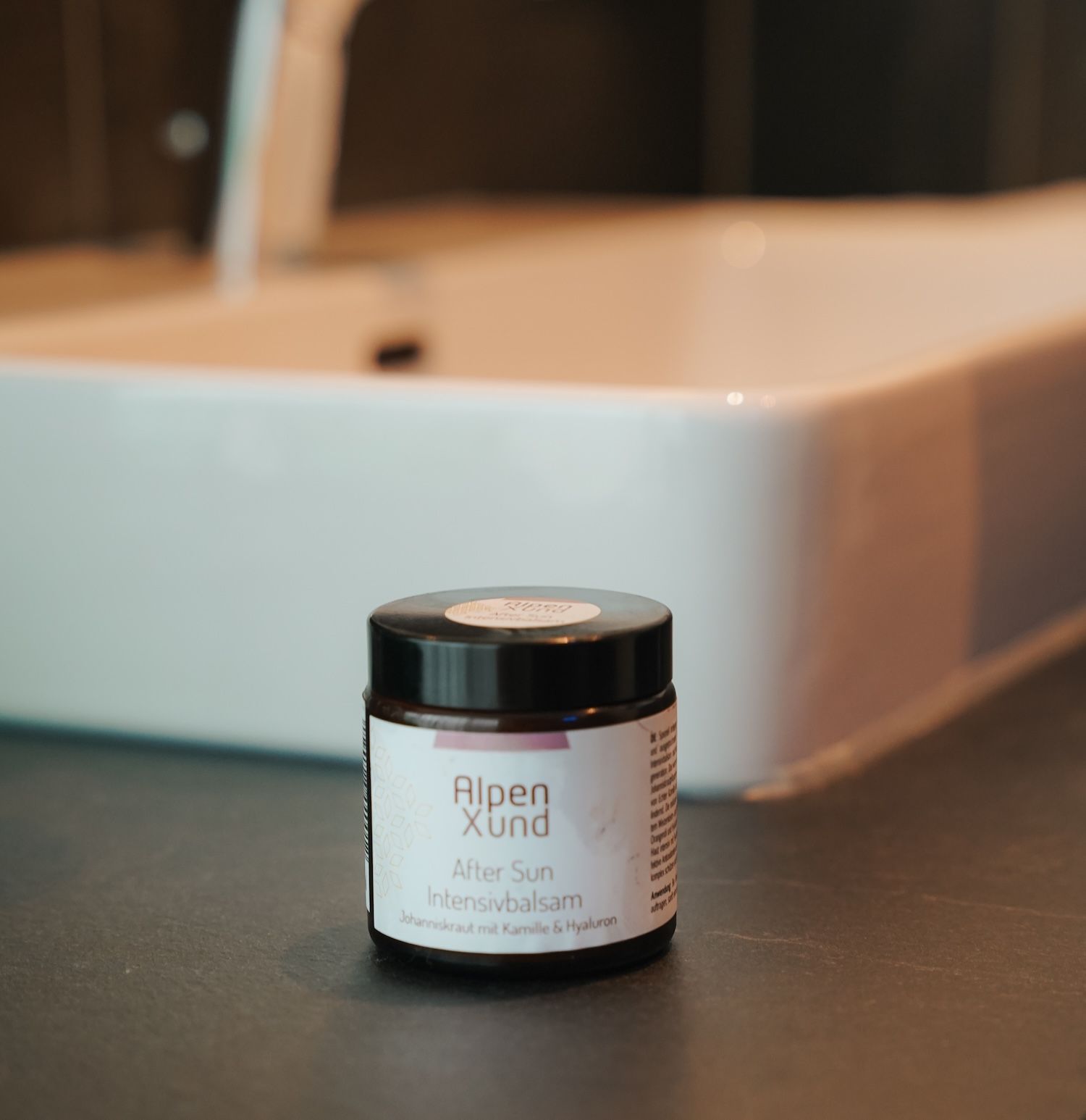 Braune Dose AlpenXund After Sun Intensivbalsam mit schwarzem Deckel steht auf einer dunklen Oberfläche. Waschbecken im Hintergrund.