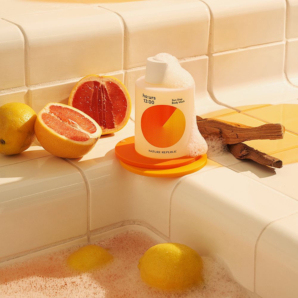 Flasche mit Schaum in einer Badewanne. Flasche: ho:urs, Sun Step Body Wash. Daneben: Obst, Holzstück.