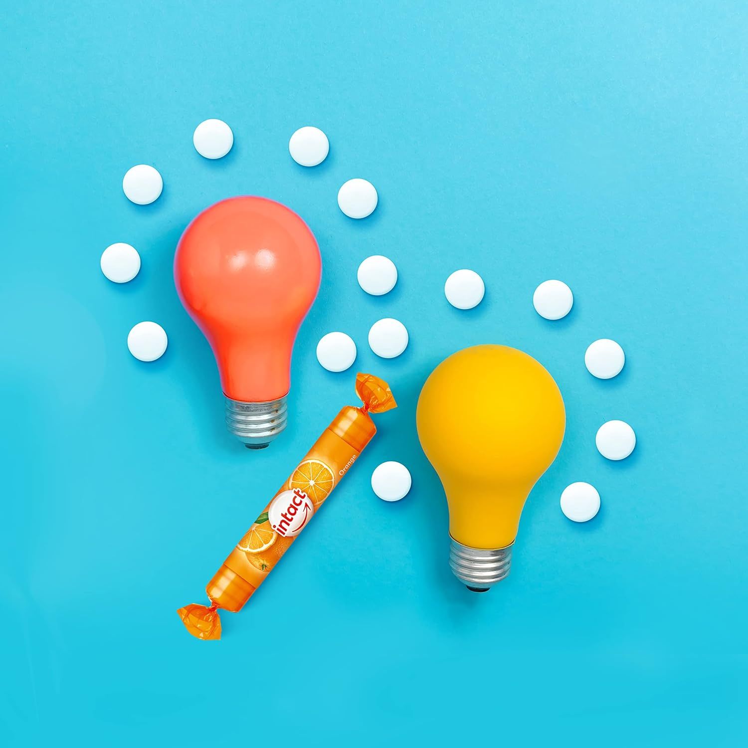 Eine Rolle Traubenzucker neben zwei Glühbirnen. Auf der Rolle ist der Schriftzug "intact" zu sehen. Daneben liegen weiße Tabletten.