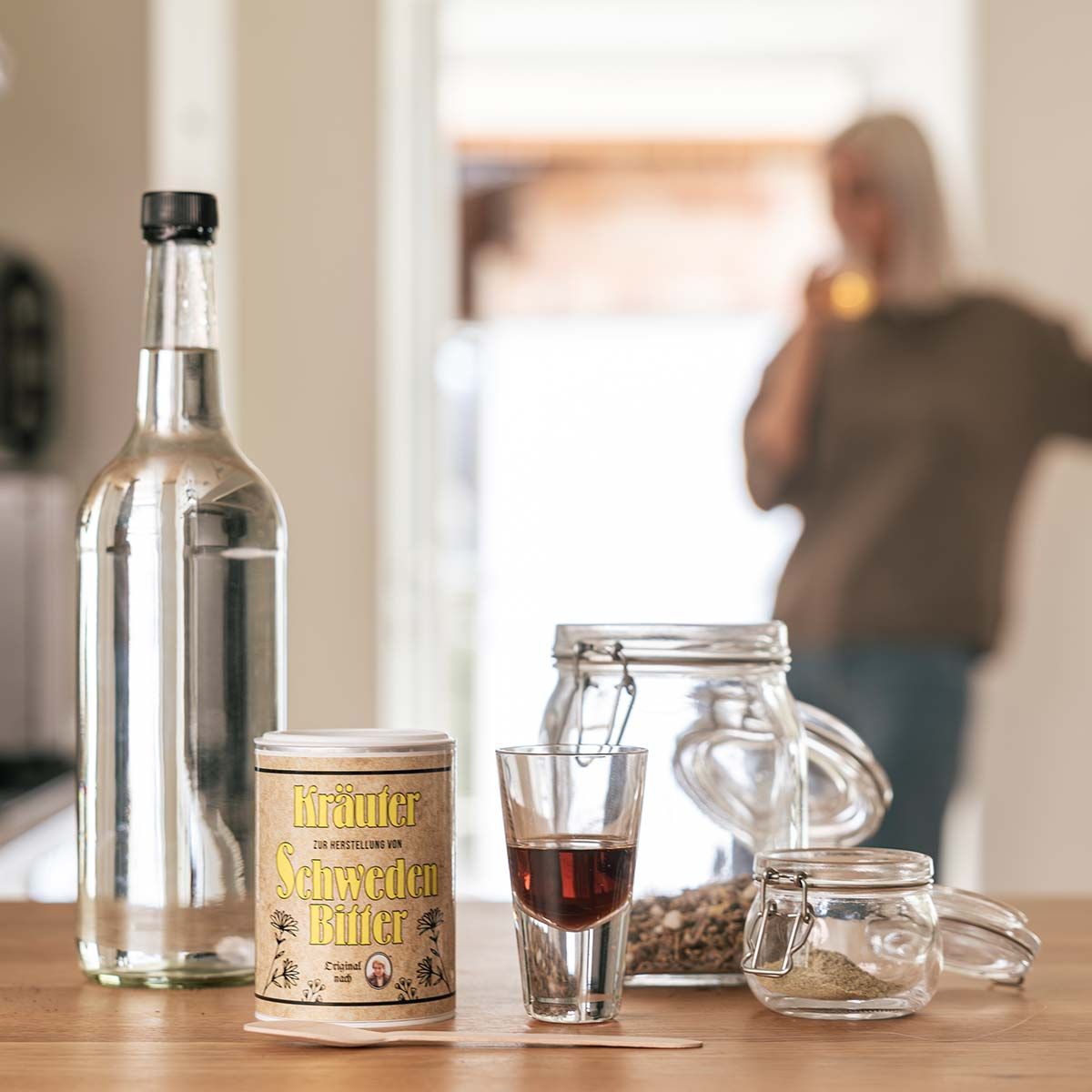 Glasflasche, Dose mit gelbem Etikett, Shotglas mit Flüssigkeit, Gläser mit Kräutern, Holzlöffel. Aufschrift: Kräuter, Schweden Bitter.