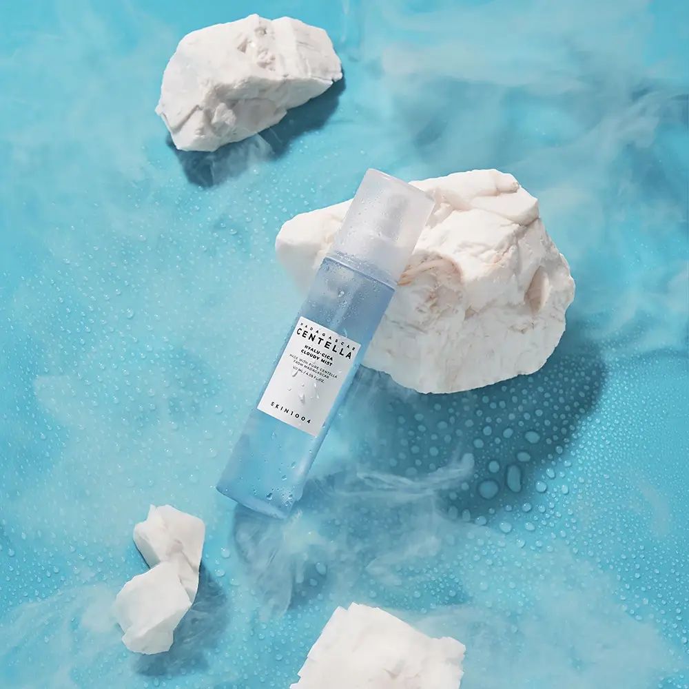 Blau-transparente Sprühflasche auf blauem Hintergrund mit weißen Steinen und Nebel. Aufschrift: MADAGASCAR CENTELLA, SKIN1004.