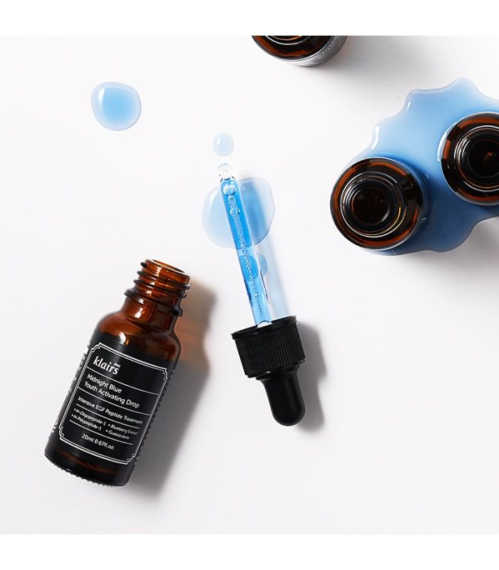 Braune Glasflasche mit schwarzem Deckel und Pipette. Blaue Flüssigkeit und Tropfen daneben. Etikett mit Text.