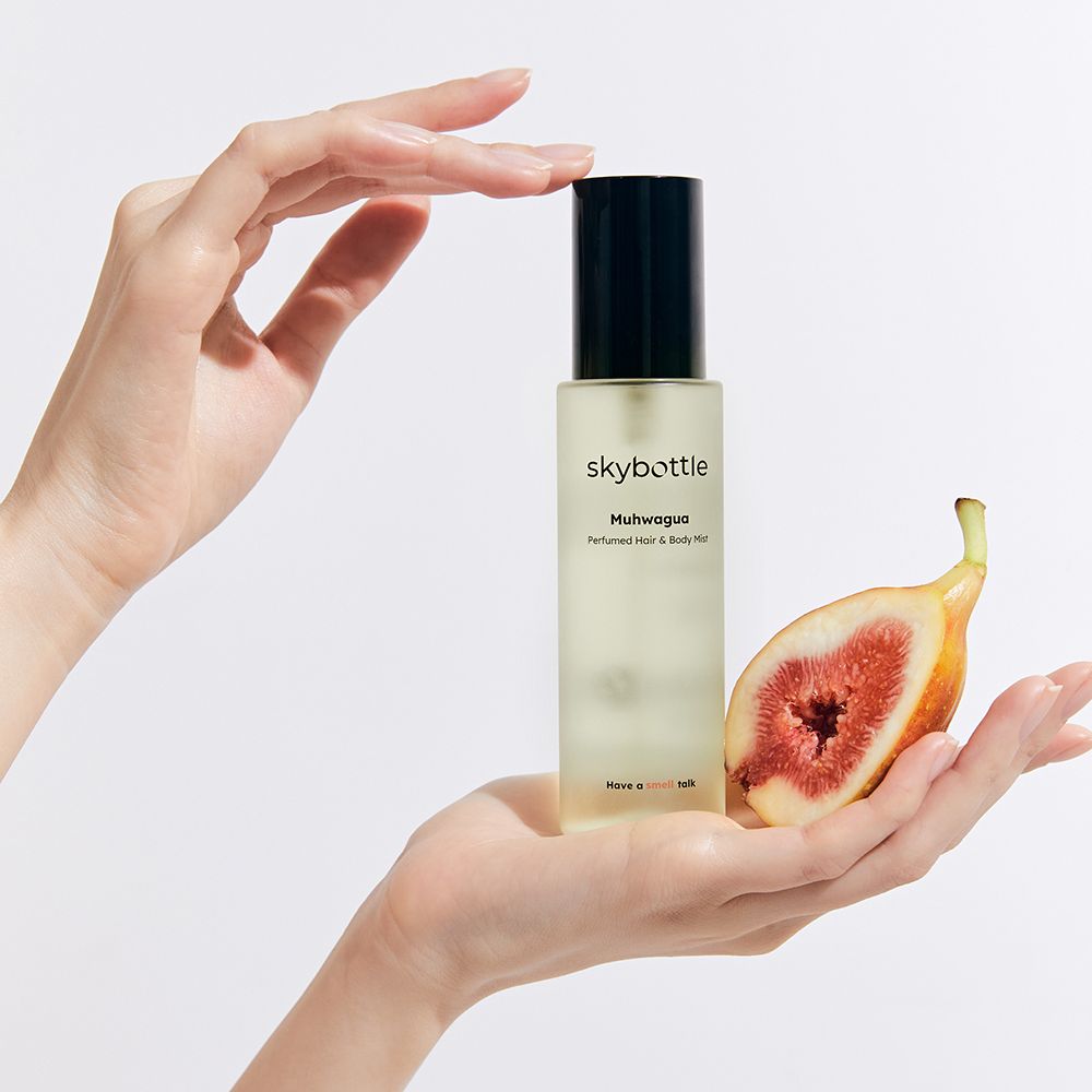 Hände halten Flakon und halbe Feige. Flakon: skybottle Muhwagua Perfumed Hair & Body Mist. Feige: rosa Fruchtfleisch, grüne Schale.