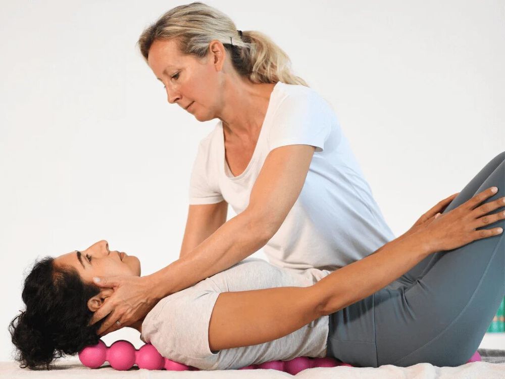 Frau liegt auf dem Rücken, Kopf auf pinkfarbenem SPINEFITTER®. Eine andere Frau massiert ihren Nacken.