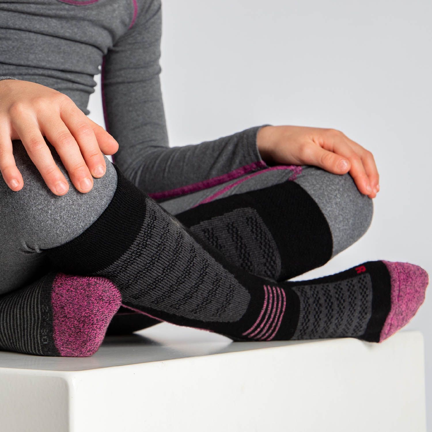 Kind sitzt mit Skisocken. Schwarze Socken mit rosa Zehen und Akzenten. Graue und schwarze Muster. Graues Outfit.