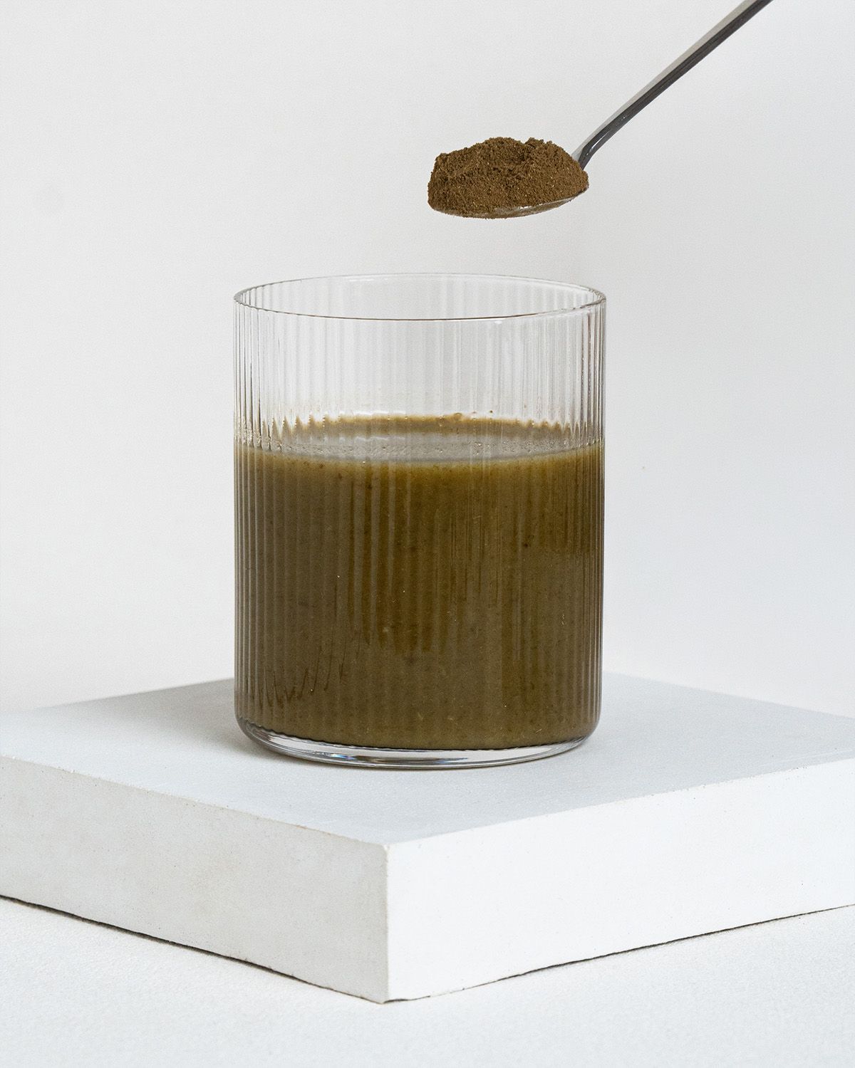 Glas mit grünem Getränk. Löffel mit braunem Pulver über dem Glas. Auf weißem Podest.