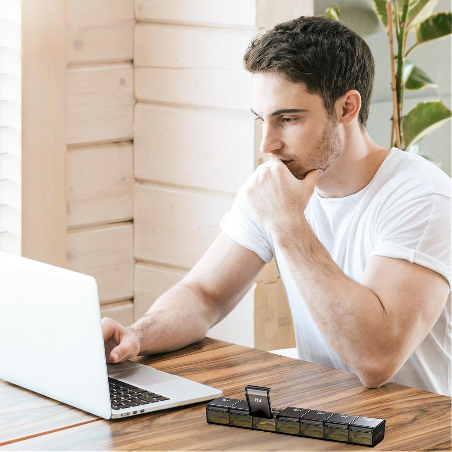 Mann sitzt am Schreibtisch und blickt auf Laptop. Pillenorganizer steht auf dem Tisch.