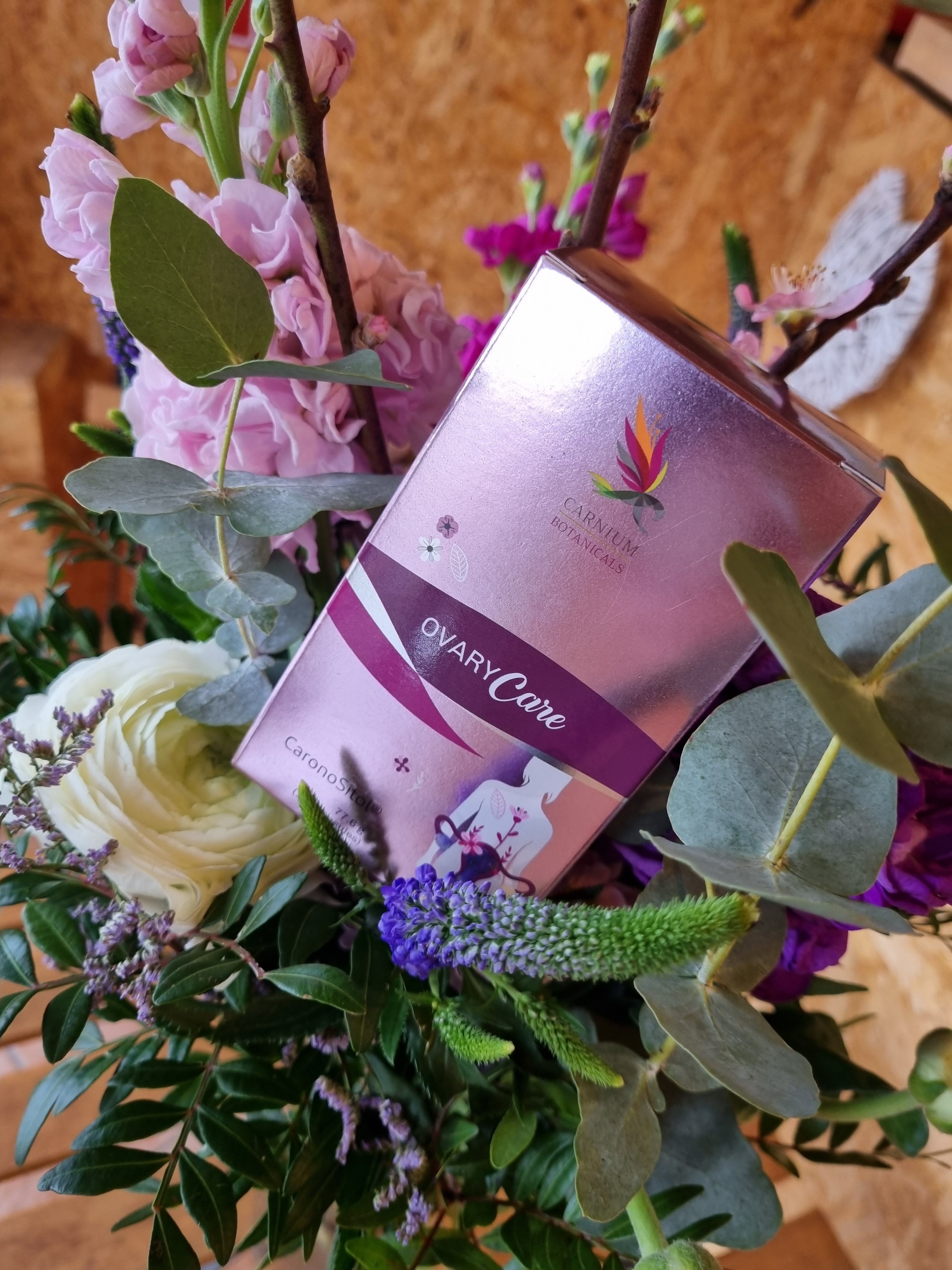 Rosa Schachtel mit Aufschrift: Carnium Botanicals Ovary Care. Umgeben von Blumen in einem Arrangement.