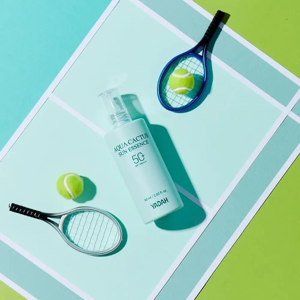 Hellgrüne Flasche mit weißem Applikator. Aufschrift: Aqua Cactus Sun Essence 50. Zwei Tennisbälle und Schläger liegen daneben.
