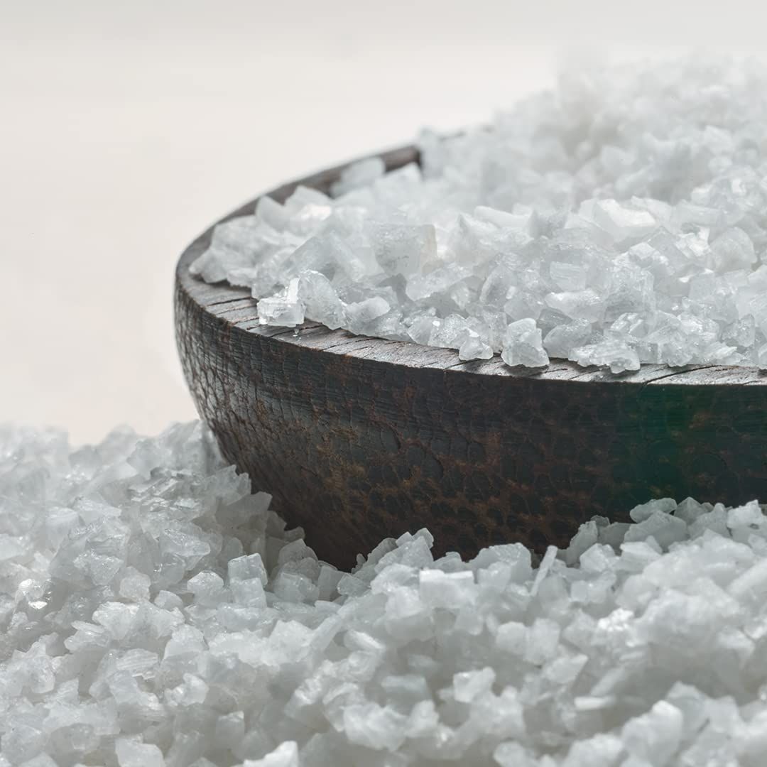 Nahaufnahme von weißen Badesalzkristallen in einer hölzernen Schale. Kristalle liegen verstreut.