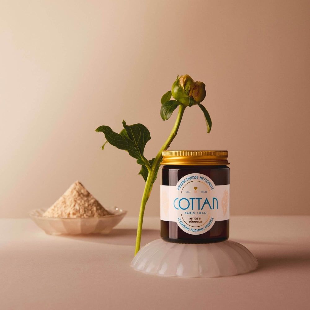 Braune Glasdose mit goldenem Deckel. Aufschrift "COTTAN". Auf einem weißen Untersetzer, daneben eine Blüte und ein Schälchen mit Pulver.