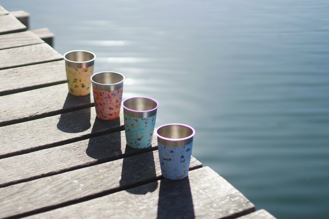 Vier bunte Becher aus Edelstahl mit Tiermotiven auf einer Holzplattform am Wasser.