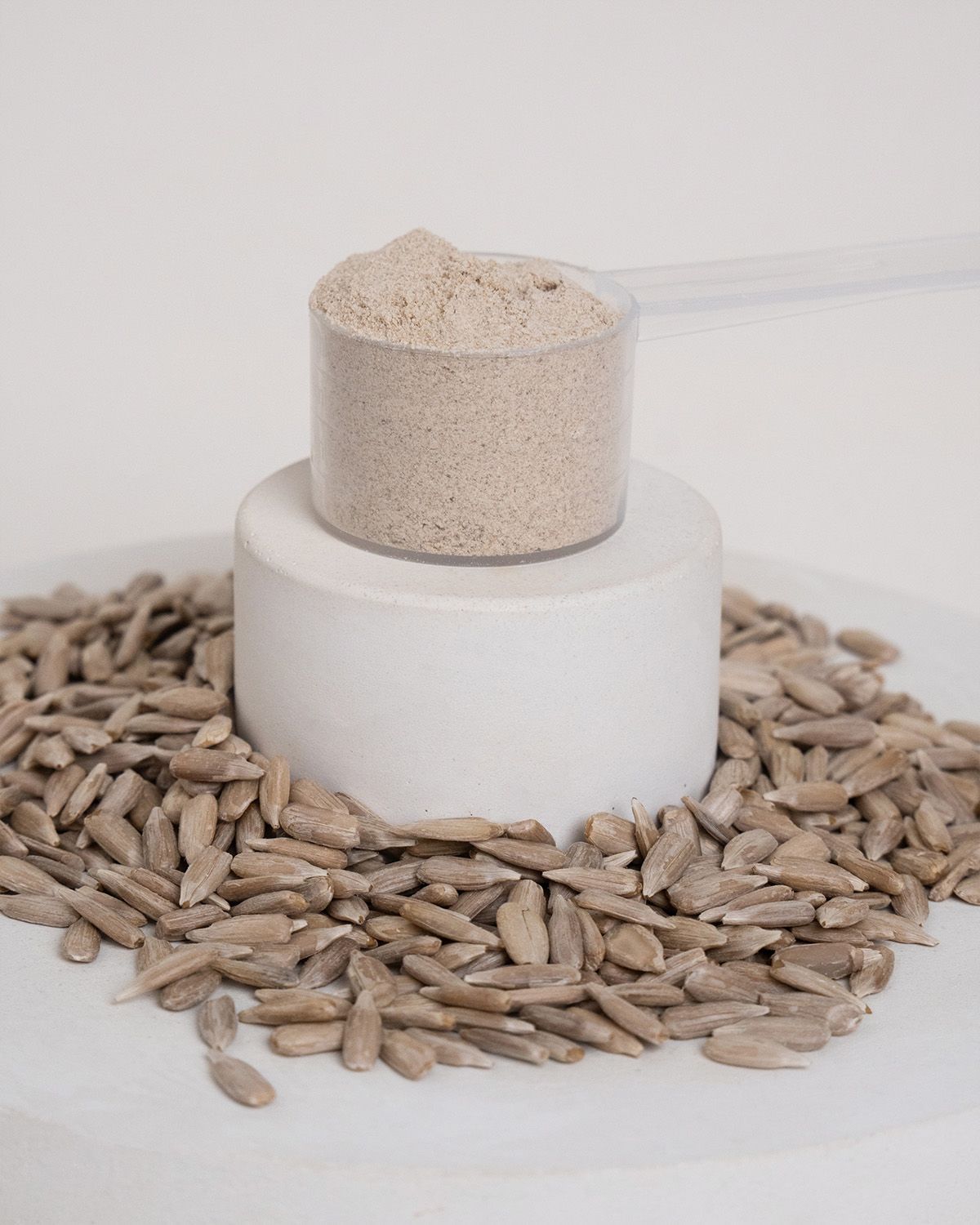 Messlöffel mit Sonnenblumenprotein-Pulver auf einem Podest, umgeben von Sonnenblumenkernen.