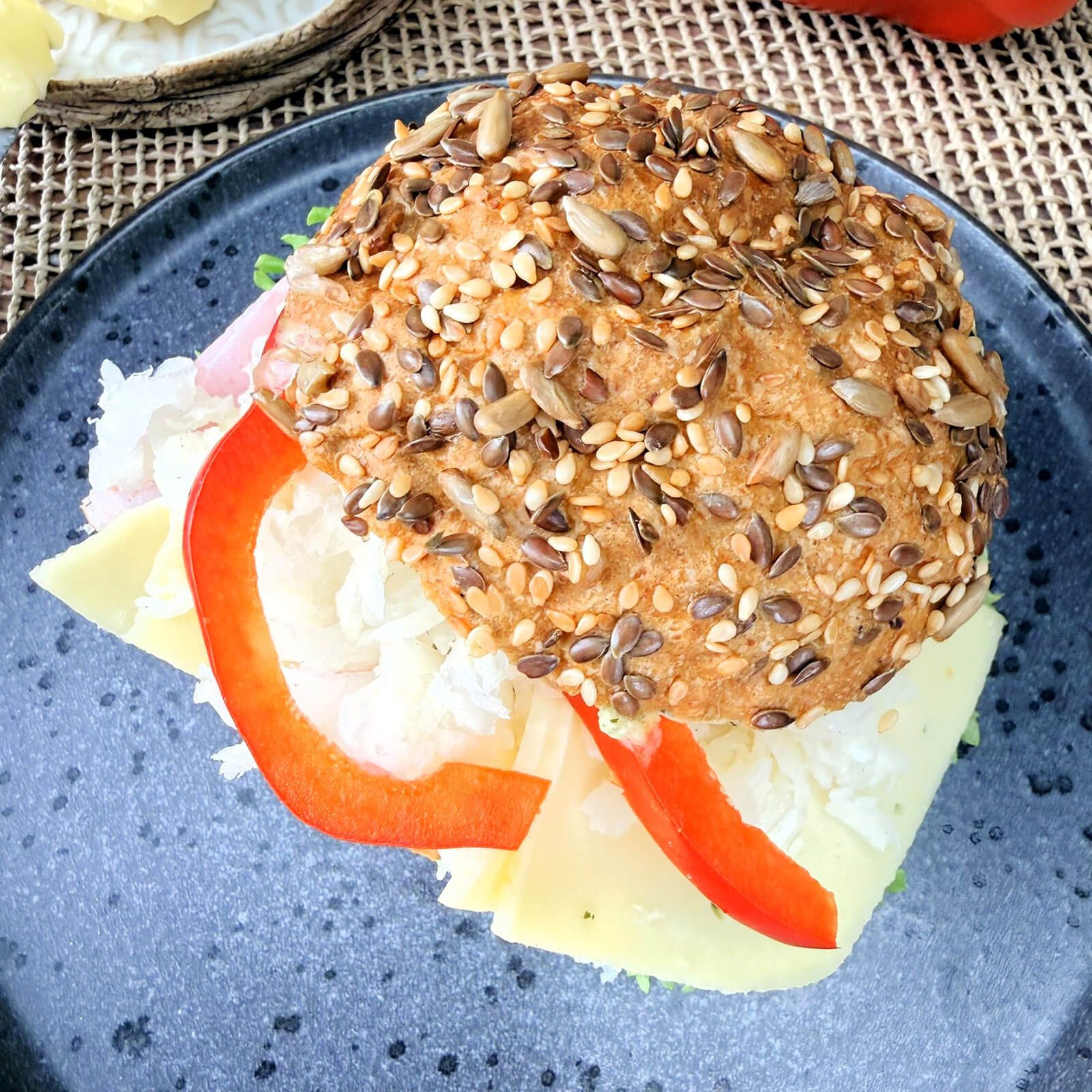 Belegtes Brötchen auf einem Teller. Brötchenhälfte mit Körnern, belegt mit Käse, Paprika und Salat. Schwarzer Teller.