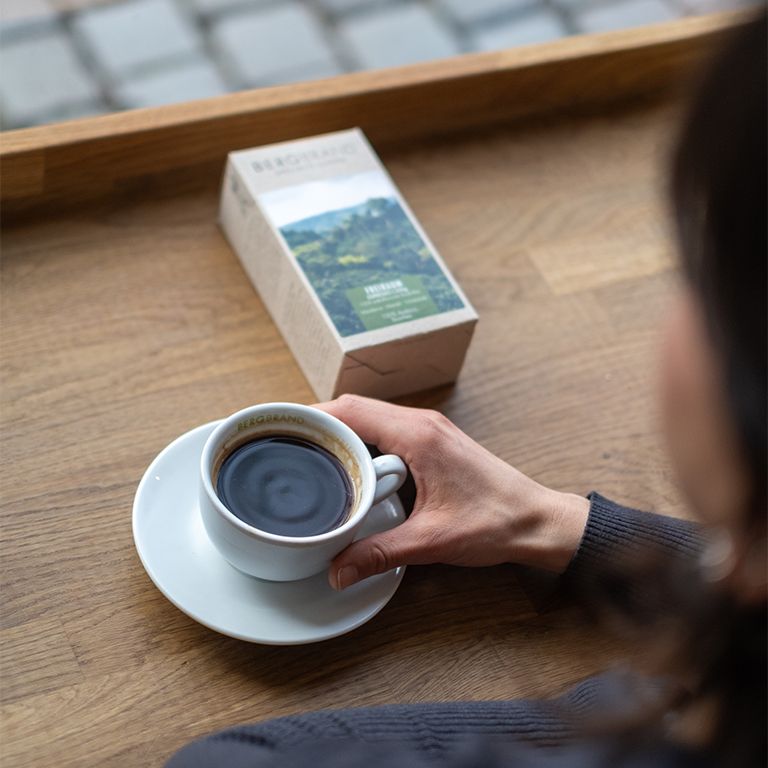 Person hält eine Tasse Kaffee auf einem Unterteller. Daneben eine Kaffeepackung. Auf einem Holztisch.